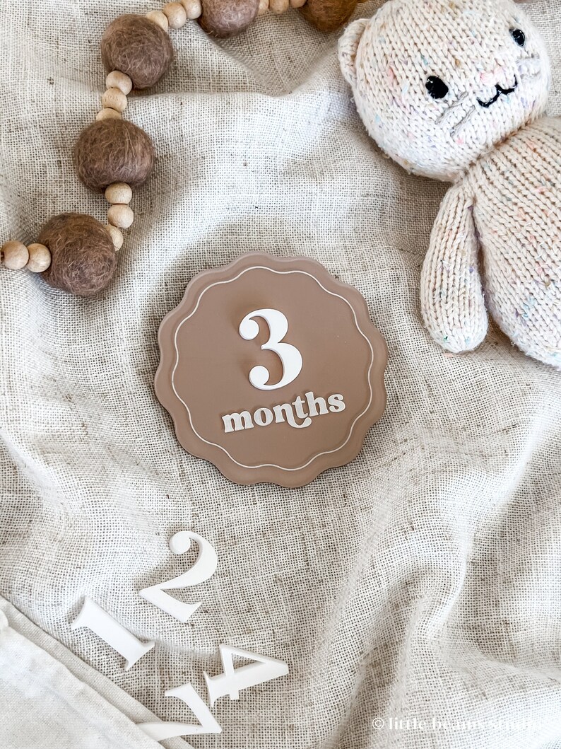 May include: A neutral-toned baby milestone marker with the number "3" and the word "months" in white script. The marker is surrounded by a scalloped edge. A knitted bear toy and wooden bead garland are also in the image, along with the numbers 1, 2, and 4.