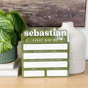 Puede incluir: Un letrero rectangular de acrílico verde oliva con texto blanco que dice "sebastian" en la parte superior, seguido de "FIRST DAY OF", "MY FAVORITE THINGS", "WHEN I GROW UP I WANT TO BE", "AGE" y "DATE" con espacios blancos para escribir.
