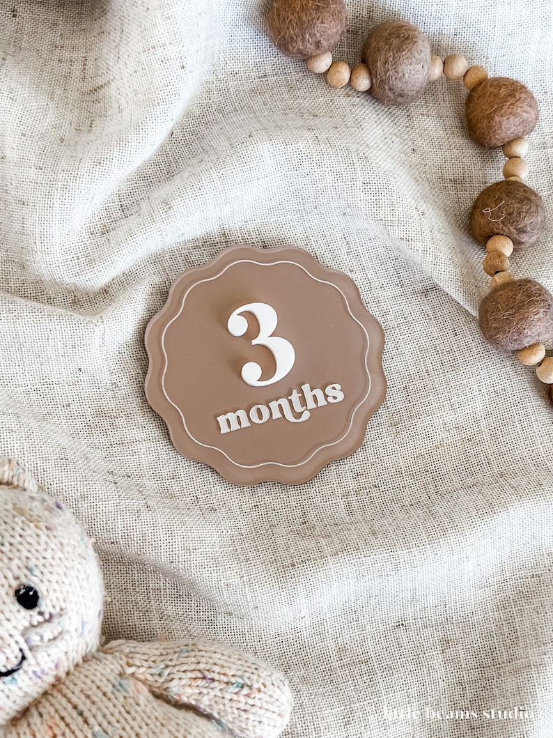 May include: A brown, scalloped-edge milestone marker with the number "3" and the word "months" in white script. The marker is on a neutral-toned fabric, with a wooden bead garland and a stuffed animal in the background.