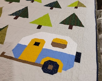 May include: A white, blue and yellow camper quilt with a quilted design of trees and a camper. The quilt is made of fabric with a white background and a blue border.