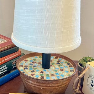 May include: A white lampshade with a black metal base sits on a rustic metal container with a mosaic tile top. The container is 6 inches in diameter. The lamp is on a wooden table with a ruler.