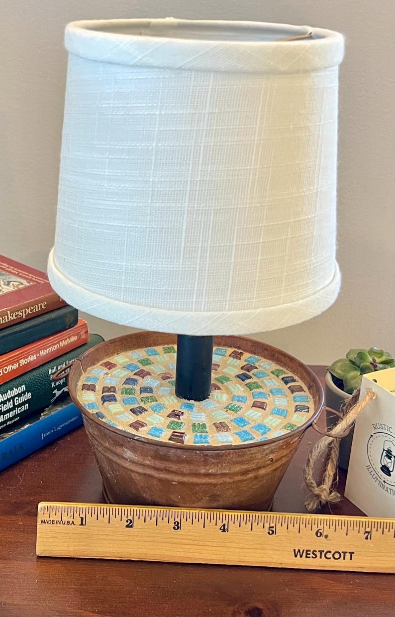 May include: A white lampshade with a black metal base sits on a rustic metal container with a mosaic tile top. The container is 6 inches in diameter. The lamp is on a wooden table with a ruler.