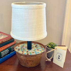 May include: A small table lamp made from a repurposed metal bucket with a mosaic tile top. The lamp has a white fabric lampshade and a black metal pole. The lamp is sitting on a wooden table with books and a small potted plant.