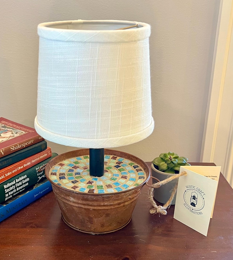 May include: A small table lamp made from a repurposed metal bucket with a mosaic tile top. The lamp has a white fabric lampshade and a black metal pole. The lamp is sitting on a wooden table with books and a small potted plant.