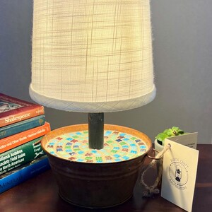 May include: A rustic lamp with a white fabric shade and a base made from a repurposed metal bucket. The bucket is decorated with a mosaic of colorful tiles. The lamp is sitting on a wooden table with a stack of books and a small potted plant.
