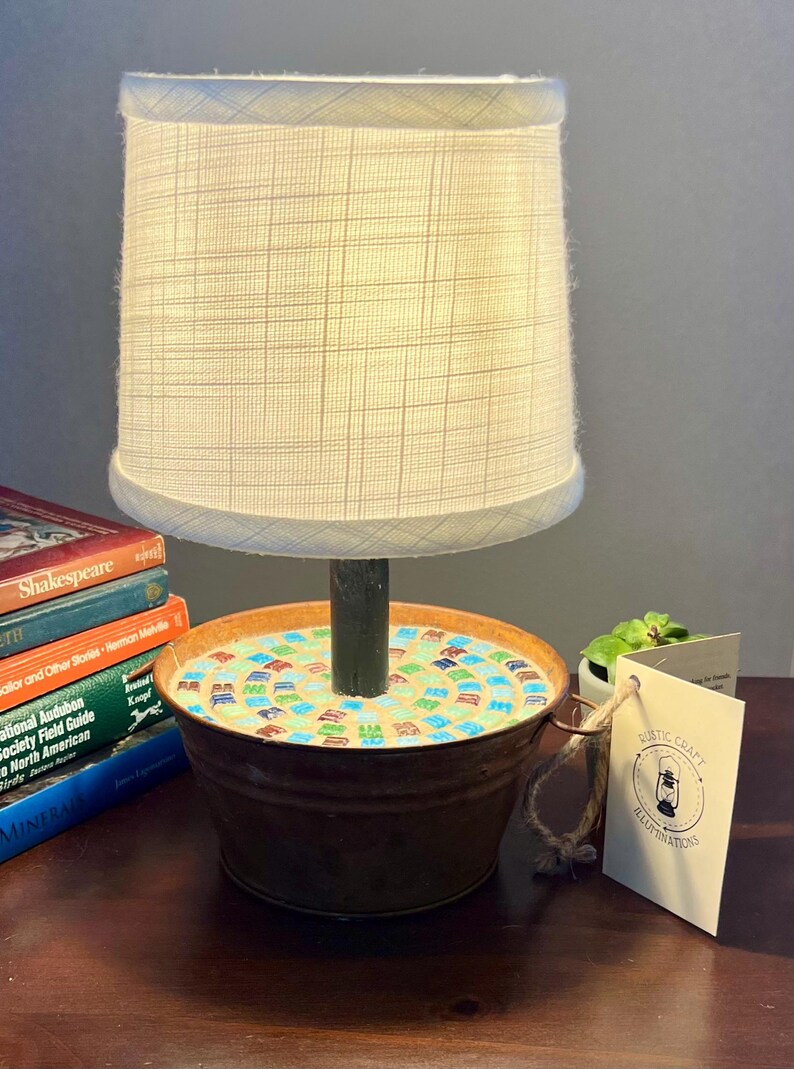 May include: A rustic lamp with a white fabric shade and a base made from a repurposed metal bucket. The bucket is decorated with a mosaic of colorful tiles. The lamp is sitting on a wooden table with a stack of books and a small potted plant.