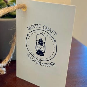 May include: A white tag with a black logo featuring a lantern inside a circle with the text "RUSTIC CRAFT ILLUMINATIONS" around it.