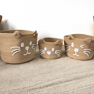 Op de afbeelding: Drie geweven beige opbergmanden met geborduurde witte kattengezichten. De manden, in verschillende maten, zijn ideaal voor het organiseren van spullen. De kattengezichten hebben snorharen en oren.