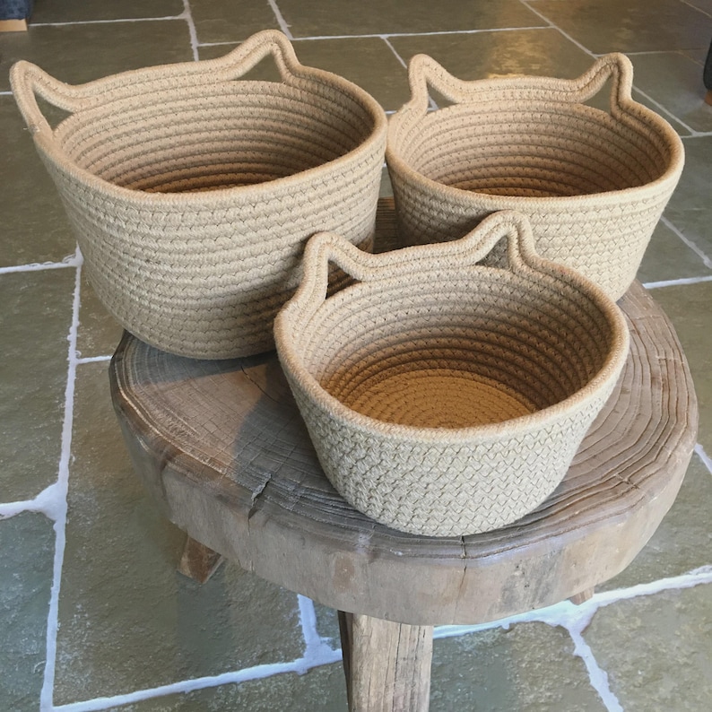 Peut inclure: Trois paniers en corde beige avec des anses en forme d'oreilles de chat sont pos&eacute;s sur une table en bois rustique. Les paniers sont de tailles diff&eacute;rentes, avec une texture tiss&eacute;e.