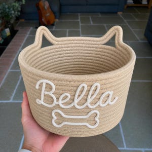 Peut inclure: Un panier en corde beige avec des anses en forme d'oreilles de chat. Le mot "Bella" est brod&eacute; en blanc en &eacute;criture cursive, avec un motif d'os blanc en dessous. Le panier est con&ccedil;u pour les fournitures pour animaux de compagnie ou le stockage.