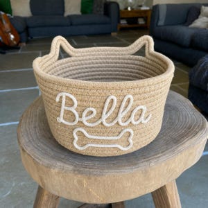 Könnte beinhalten: Ein beiger Seilkorb mit ohrenförmigen Griffen. Der Name "Bella" ist in weißer Schreibschrift gestickt, darunter eine weiße Knochengrafik. Der Korb steht auf einem Holzschemel.