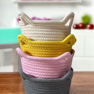 Peut inclure: Quatre paniers en corde empil&eacute;s, de couleurs cr&egrave;me, jaune, rose et grise. Chaque panier a deux poign&eacute;es. Les paniers sont pos&eacute;s sur une surface en bois, avec un arri&egrave;re-plan flou d'une &eacute;tag&egrave;re blanche et d'objets rouges et violets.