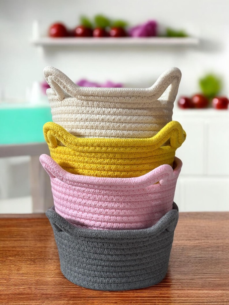 Peut inclure: Quatre paniers en corde empil&eacute;s, de couleurs cr&egrave;me, jaune, rose et grise. Chaque panier a deux poign&eacute;es. Les paniers sont pos&eacute;s sur une surface en bois, avec un arri&egrave;re-plan flou d'une &eacute;tag&egrave;re blanche et d'objets rouges et violets.