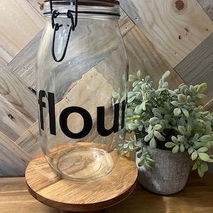 May include: A clear glass jar with a black label that says "flour" on it. The jar has a metal clasp and is sitting on a wooden stand. A small potted plant is next to the jar.