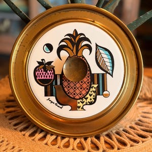 May include: A round brass tray featuring a central white ceramic inlay with a stylized pineapple graphic in orange, black, and gold. The design includes a small strawberry, a leaf, and the signature Georges Briand in black script on the white surface.