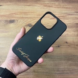 Op de afbeelding: Zwarte telefoonhoes met een gouden Apple-logo en de naam "Scarlet Thongson" in gouden letters. De hoes wordt in een hand gehouden tegen een houten achtergrond.