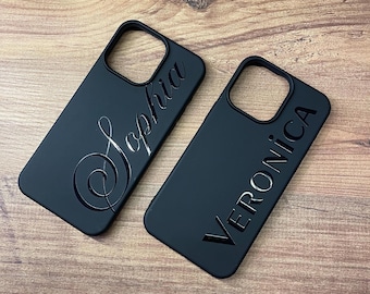 May include: Two black phone cases with personalized names. One case says "Sophia" in a cursive font, and the other says "Veronica" in a sans-serif font. The cases are on a wooden surface.