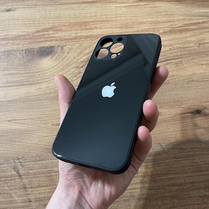 May include: A sleek black phone case with a glossy surface. It has cutouts for the camera and a white Apple logo. The case is designed to protect the phone from scratches and other damage.
