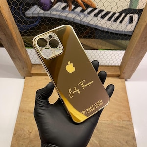Op de afbeelding: Een gouden iPhone-hoes met een 24KT Gold Luxury Edition-opschrift. De hoes heeft een transparante achterkant en een zwarte camera-lensafdekking. De naam "Emily Thompson" is gegraveerd op de achterkant van de hoes.
