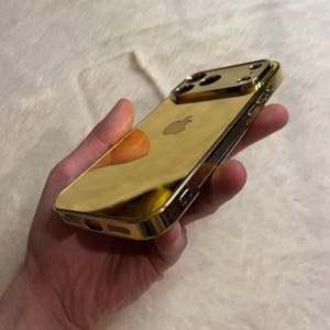 Op de afbeelding: Een goudkleurige telefoonhoes met een reflecterend oppervlak. De hoes heeft een strak ontwerp met uitsparingen voor de camera en knoppen. Het Apple-logo is zichtbaar op de achterkant. De hoes wordt in een hand gehouden.