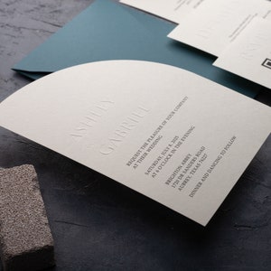 Puede incluir: Una invitación de boda blanca con la parte superior curvada y texto en relieve. La invitación dice "Ashley & Gabriel solicitan el placer de su compañía en su boda el sábado 8 de julio de 2025 a las 6 en punto de la tarde Brighton Abbey 1720 Dr Sanders Road Aubrey, Texas 76227 Cena y baile a continuación."