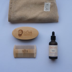 Puede incluir: Un conjunto de productos para el cuidado de la barba que incluye un peine de madera, un cepillo de madera, una botella marrón de aceite para barba y una toallita beige. Los productos están etiquetados con el texto "DEVA Beard Grooming" y "देवा".