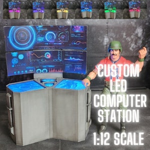 Puede incluir: Una estación de ordenador LED personalizada a escala 1:12, con un diseño gris de tres piezas y luces LED azules. La estación tiene dos monitores con pantallas azules brillantes que muestran información digital. Una figura de acción a escala 1:12 se encuentra junto a la estación.