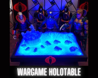 Può includere: Un tavolo nero con una superficie illuminata di blu e un logo Cobra. Il tavolo è circondato da action figure, tra cui una figura di Cobra Commander. Il testo "WARGAME HOLOTABLE" è stampato sul tavolo.