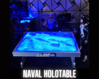 Può includere: Un tavolo in plastica blu illuminato dal basso, con una cornice bianca e le parole "GI JOE" stampate sul davanti. Il tavolo è illuminato dal basso, creando un bagliore blu. Il tavolo è un modello di una scena di battaglia navale, con navi e isole visibili nel bagliore blu.