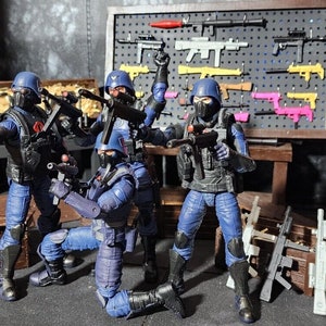 Puede incluir: Figuras de acción de cuatro soldados con uniformes y cascos azules, armados con rifles y pistolas. Una exhibición de armas de juguete está montada en la pared detrás de ellos, con armas adicionales en una mesa y en una caja de madera.