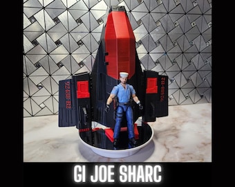 May include: A black and red toy jet with a figure of a man in a blue uniform standing in front of it. The jet has the text "92488-B2" on the side. The toy is on a black and white display stand. GI JOE SHARC is written in white letters on a black background.