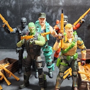 Puede incluir: Figuras de acción de varios personajes, cada uno sosteniendo un rifle. Las figuras visten atuendos de estilo militar, con una variedad de colores que incluyen verde, negro y beige. Cajas de madera con rifles en primer plano.