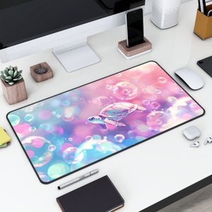May include: A pink and blue mouse pad with a sea turtle swimming through iridescent bubbles. The mouse pad is on a white desk with a computer monitor, a cell phone, a small white speaker, and a black and white mouse.