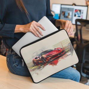 May include: A laptop sleeve with a red sports car illustration on a beige background. The car is a Ferrari with a black interior and black wheels. The sleeve has a black zipper.