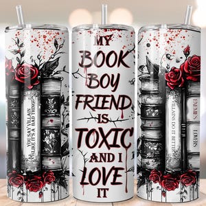 May include: A white tumbler with a straw, featuring a design of stacked books, red roses, and the text "MY BOOK BOYFRIEND IS TOXIC AND I LOVE IT." The design includes the phrases "YOU SAY VILLAIN LIKE IT'S A BAD THING" and "VILLAINS DO IT BETTER."