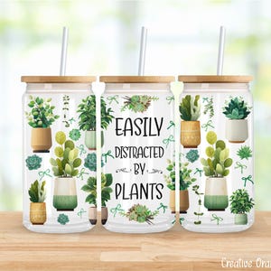 Puede incluir: Vaso de vidrio transparente con tapa y pajita de bambú, decorado con plantas en macetas y el texto "EASILY DISTRACTED BY PLANTS". El diseño presenta varias plantas verdes y suculentas en macetas.