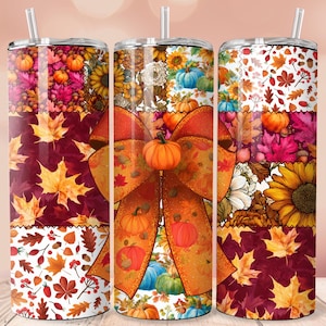 May include: Three stainless steel tumblers with fall-themed designs. Each tumbler features a patchwork design with pumpkins, sunflowers, and autumn leaves in shades of orange, red, and yellow. A large orange bow with a pumpkin center adorns the center tumbler.
