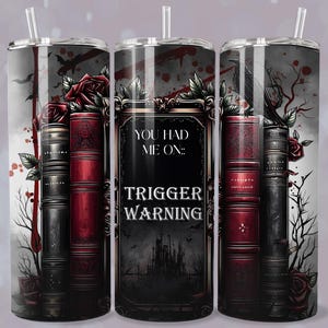 May include: A stainless steel tumbler with a gothic design. The tumbler features a stack of books, roses, and the text "YOU HAD ME ON: TRIGGER WARNING." The design is in shades of red, black, and gray, with a straw.