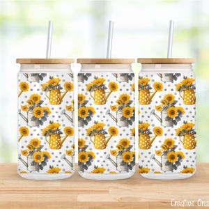Puede incluir: Tres vasos de vidrio con tapas y pajitas de bambú. Los vasos presentan un patrón repetido de girasoles, regaderas amarillas y lunares grises. El fondo es una imagen borrosa de una superficie de madera y un fondo verde y blanco.