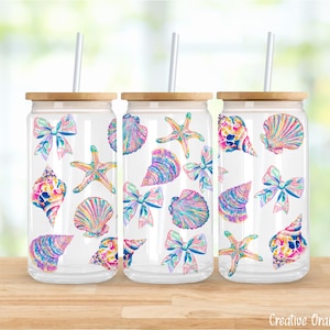 Puede incluir: Tres vasos de vidrio transparente con tapas de bambú y pajitas blancas. Cada vaso está decorado con ilustraciones coloridas de conchas marinas y lazos. Los vasos están sobre una superficie de madera con un fondo borroso.