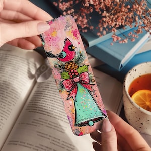 May include: A colorful bookmark featuring a red cardinal perched on a pinecone and holly. Below the bird is a teal bell with a red bow. The background has a pink and yellow watercolor effect. The bookmark is held over an open book.