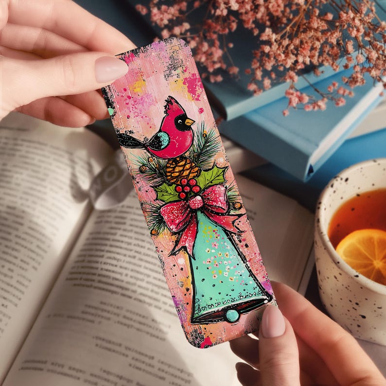 May include: A colorful bookmark featuring a red cardinal perched on a pinecone and holly. Below the bird is a teal bell with a red bow. The background has a pink and yellow watercolor effect. The bookmark is held over an open book.