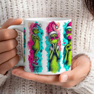 May include: White ceramic mug with a Grinch design in different poses, wearing a Santa hat and heart-shaped sunglasses. The Grinch is green with pink and teal accents, and small hearts. The mug has a white handle.