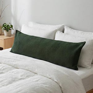 May include: A long, dark green corduroy body pillow rests on a bed with white linens and pillows. The pillow is rectangular and has a ribbed texture. A wooden nightstand with a vase and a small plant is visible in the background.