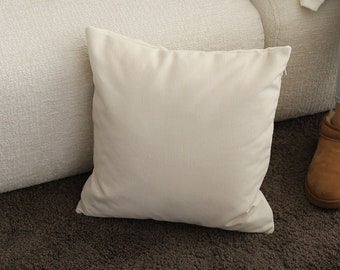 May include: Off-white rectangular and square throw pillows on a cream-colored sofa. A dark wooden tray with a cup and an open book sits on the sofa. The pillows are made of a soft, textured fabric.
