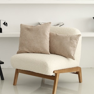 May include: A white armchair with a fluffy, sheepskin-like upholstery. Two beige throw pillows are placed on the seat. The chair has a wooden frame and legs. The chair is in front of a white shelf with various decorative objects.