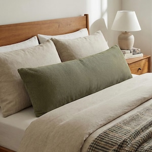 May include: A bed with a wooden headboard, featuring a long, olive green corduroy pillow. The bed is made with beige bedding and a plaid throw blanket. A lamp sits on a wooden nightstand.
