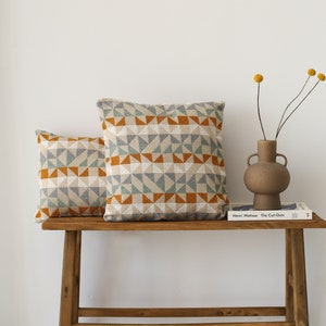 Op de afbeelding: Twee gepatroneerde kussens in beige, oranje en blauwe tinten liggen op een houten bank. Een boek getiteld "Henri Matisse: The Cut-Outs" ligt op de bank. Een vaas met gedroogde gele bloemen staat op de bank.