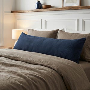 Könnte beinhalten: Ein marineblaues Kord-Körperkissen liegt auf einem Bett mit beigefarbener Bettwäsche. Das lange, rechteckige Kissen hat vertikale Rippen. Zwei beige Kissen befinden sich hinter dem Körperkissen. Eine Lampe, Kunstwerke und Dekorationsartikel befinden sich auf einem Regal.