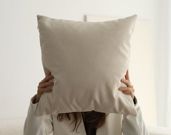 May include: A person sitting on a white couch, holding a large, off-white pillow in front of their face.